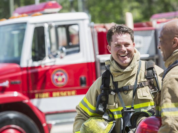 Two firefighters in gear are smiling near a bright red fire truck outdoors, appearing to have a casual conversation.