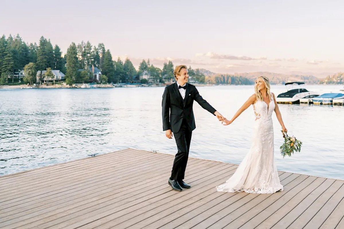 A couple in wedding attire holds hands on a dock by the water at sunset.