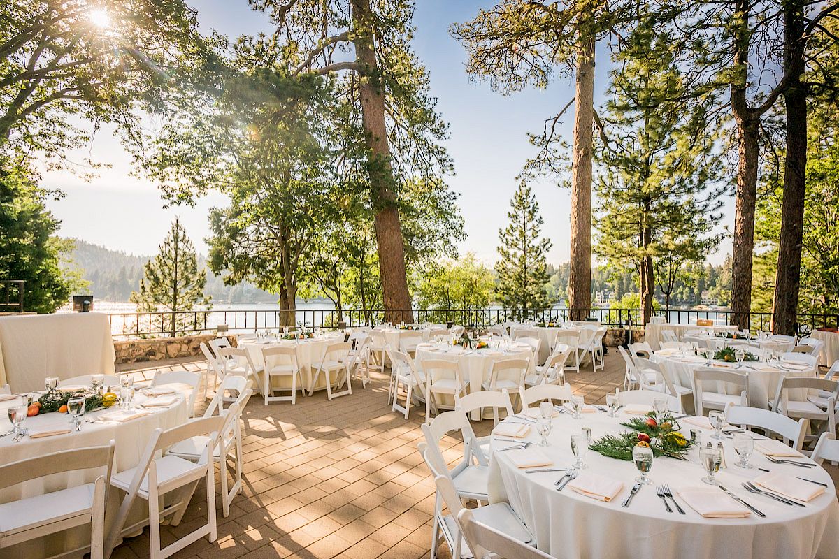 Outdoor event setting with white chairs and tables, near trees with a lake view.