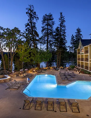 A hotel pool deck by a lake at dusk, with lounge chairs, trees, and a multi-story building under a clear blue sky.
