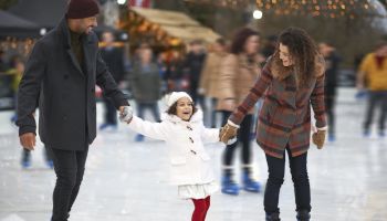 A family is ice skating together, holding hands and smiling, with other skaters and festive lights in the background.