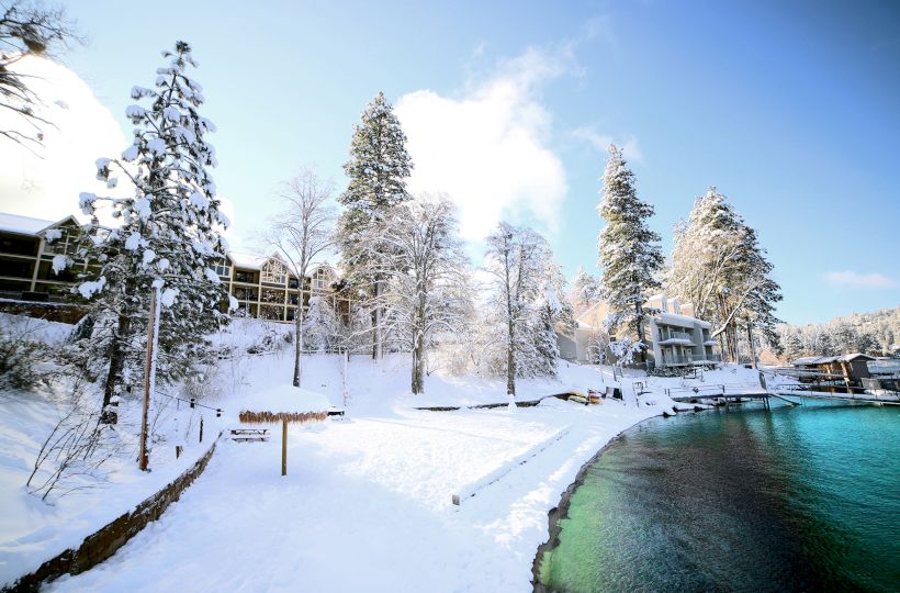 A snowy landscape with trees and buildings by a clear lake under a bright blue sky.