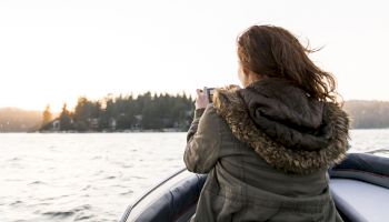 A person in a jacket is on a boat taking a photo of a scenic view with trees and water during sunset.