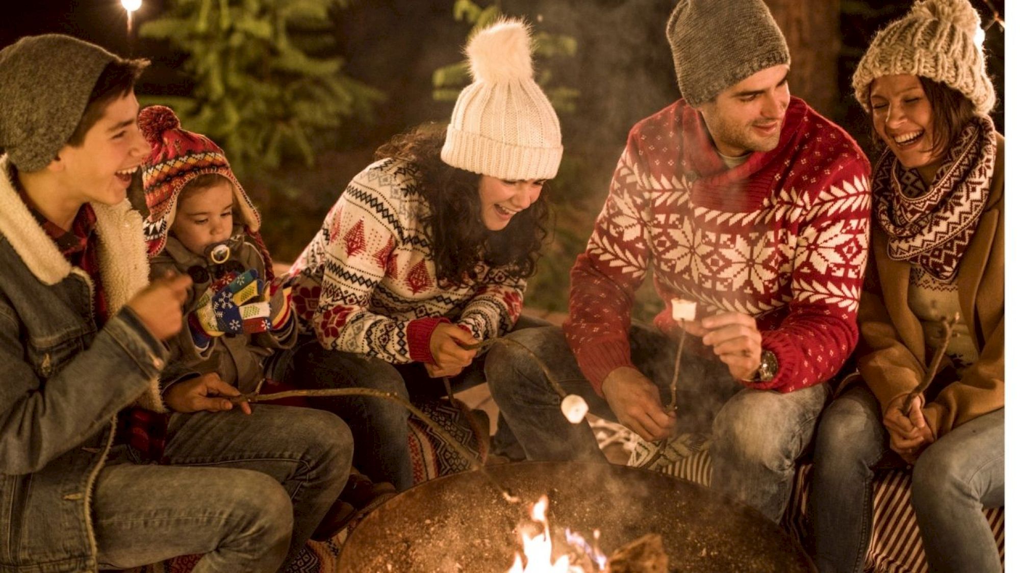 A group of people in winter clothing are sitting around a campfire, roasting marshmallows, and enjoying each other's company outdoors.