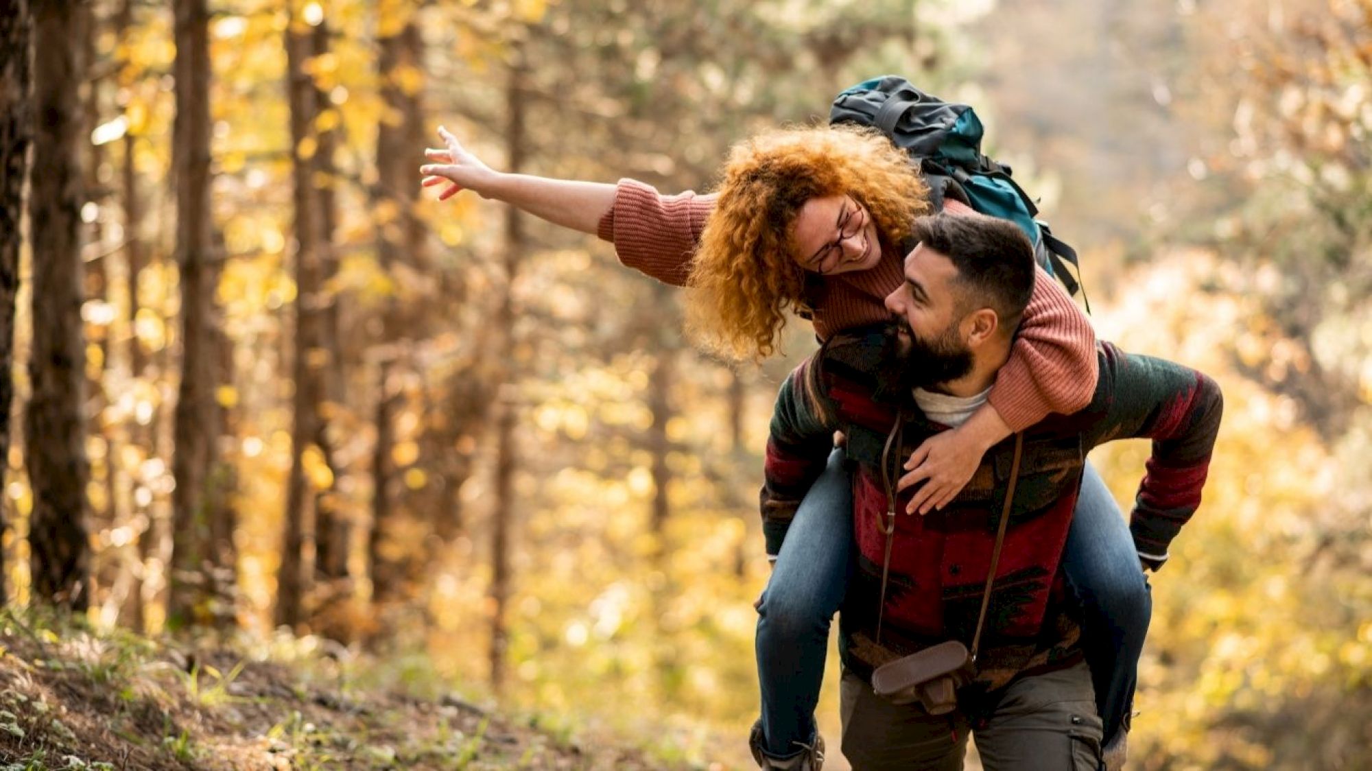 A person gives another a piggyback ride through a forest. Their expressions show happiness, surrounded by autumn trees.