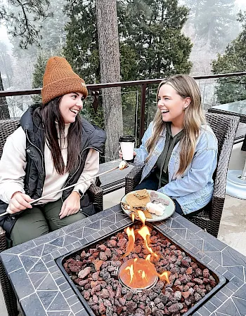 Two friends in winter jackets laugh by a glowing outdoor firepit, sipping coffee and sharing food on a terrace.